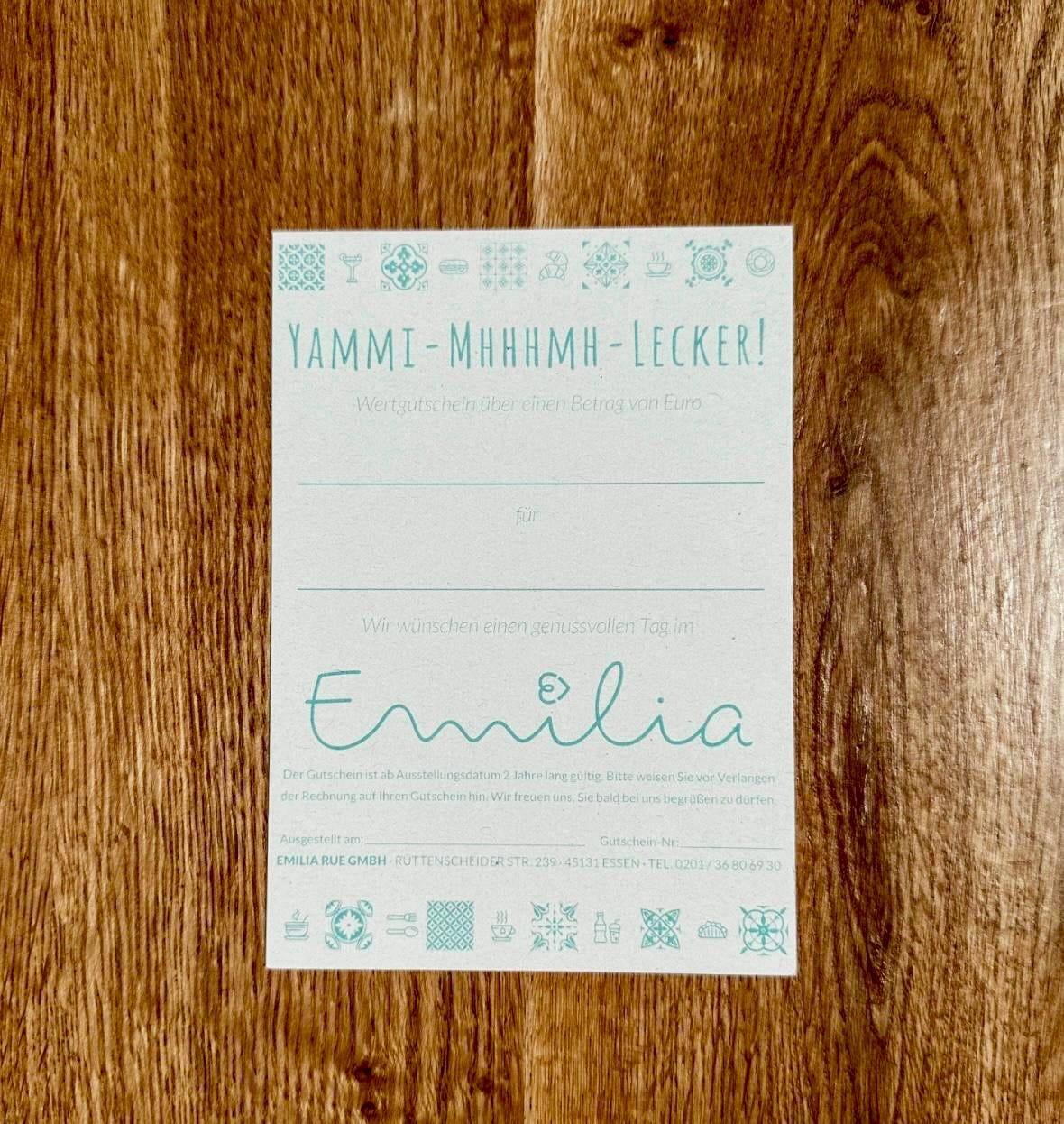 Ein Gutschein des Emilia auf einem Holztisch, daneben eine Tasse Kaffee und ein frischer Blumenstrauß – perfekt als Geschenk zum Muttertag.
