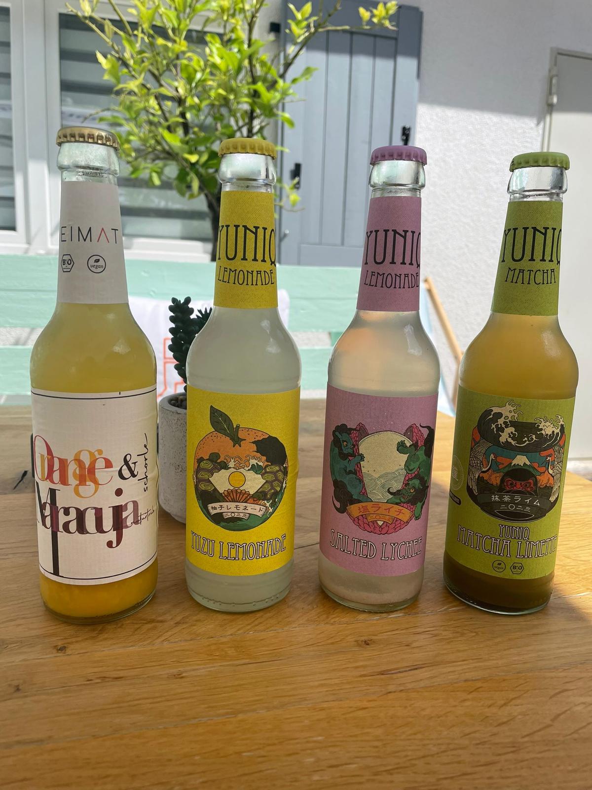 Vier bunte Glasflaschen mit Bio-Limonade auf einem Holztisch im Sonnenschein. Sorten: HEIMAT Orange & Maracuja, YUNIQ Yuzu Lemonade, YUNIQ Salted Lychee, YUNIQ Matcha Limette. Im Hintergrund grüne Pflanzen und hellblaue Fensterläden – sommerliche Terrassenstimmung im Cafe Emilia Rue.